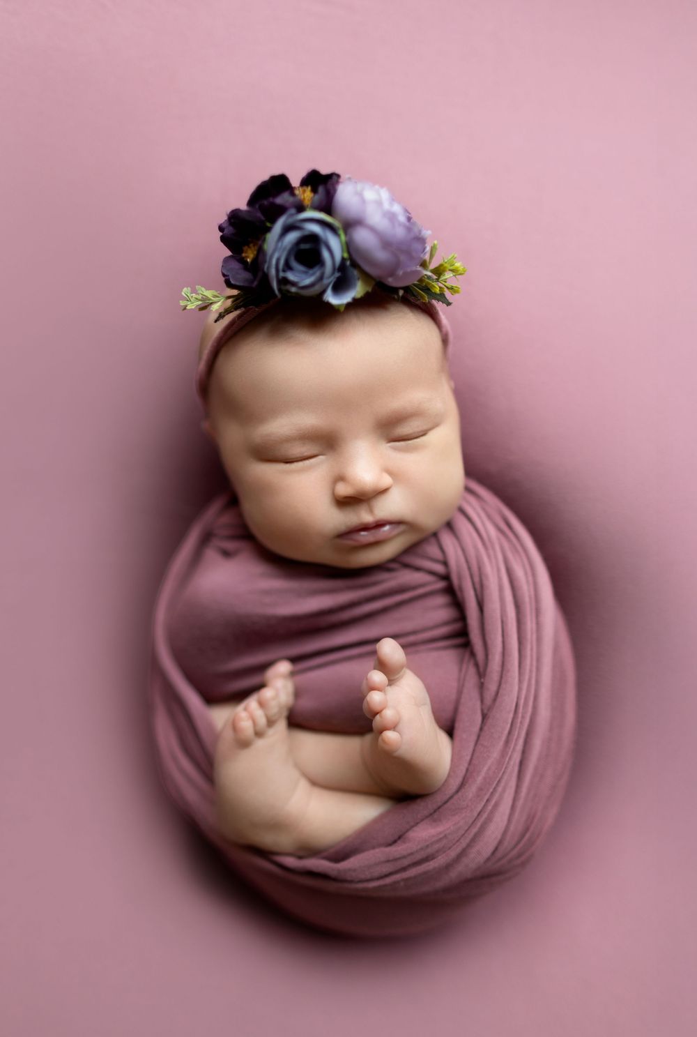Sleeping newborn wrapped in soft mauve fabric with purple floral headband on pink background.