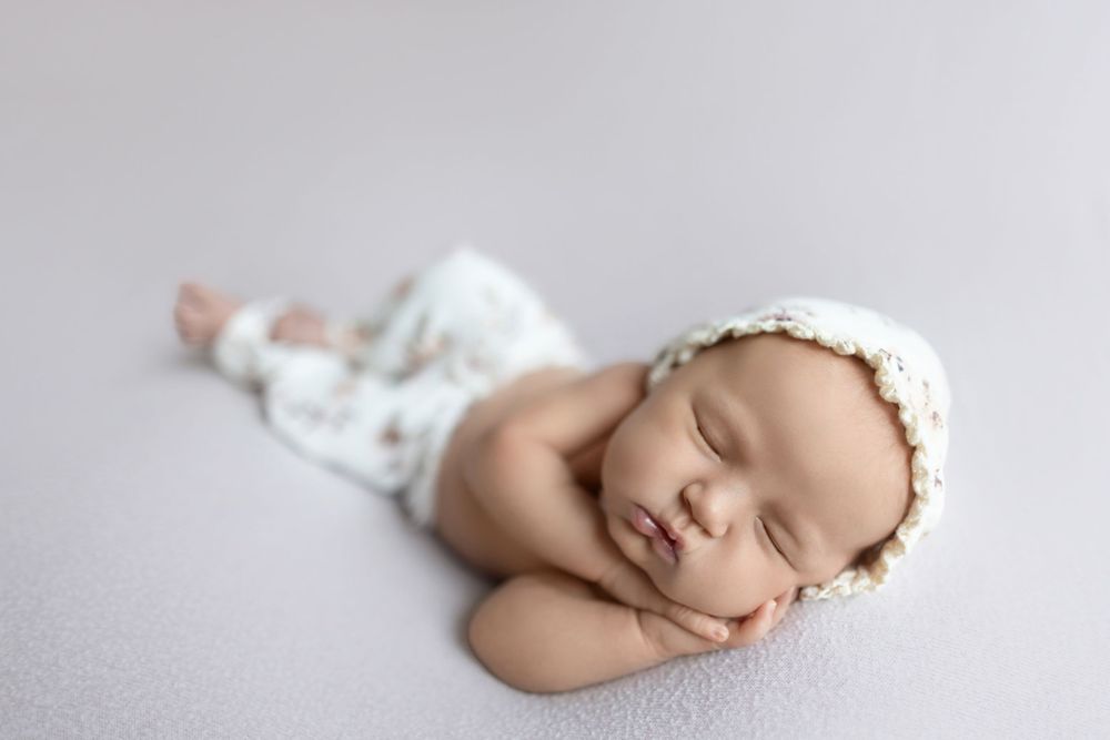 Peaceful sleeping newborn baby wearing white bonnet and pants rests on soft gray backdrop during professional photo session.