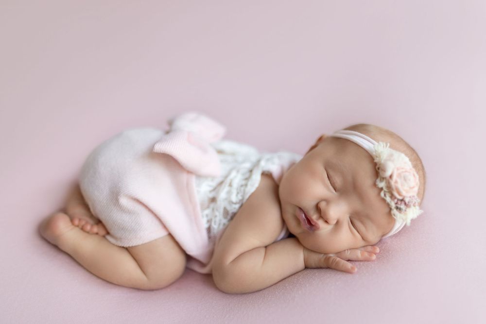 A sleeping newborn baby wearing white clothing lies peacefully on a soft pink blanket in a studio setting.