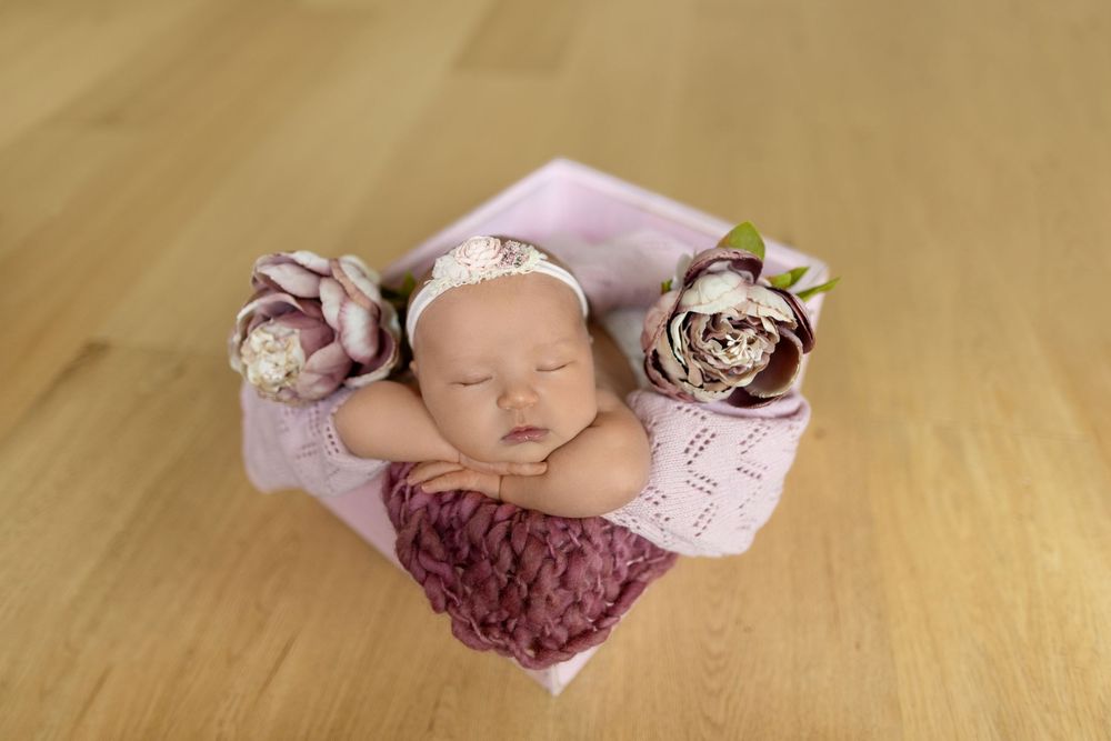A sleeping newborn baby wearing pink and burgundy accessories rests peacefully on a wooden floor.