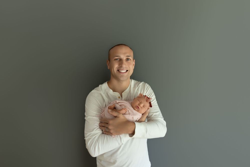 A person in a white sweater holds a small newborn baby while smiling warmly against a gray background.