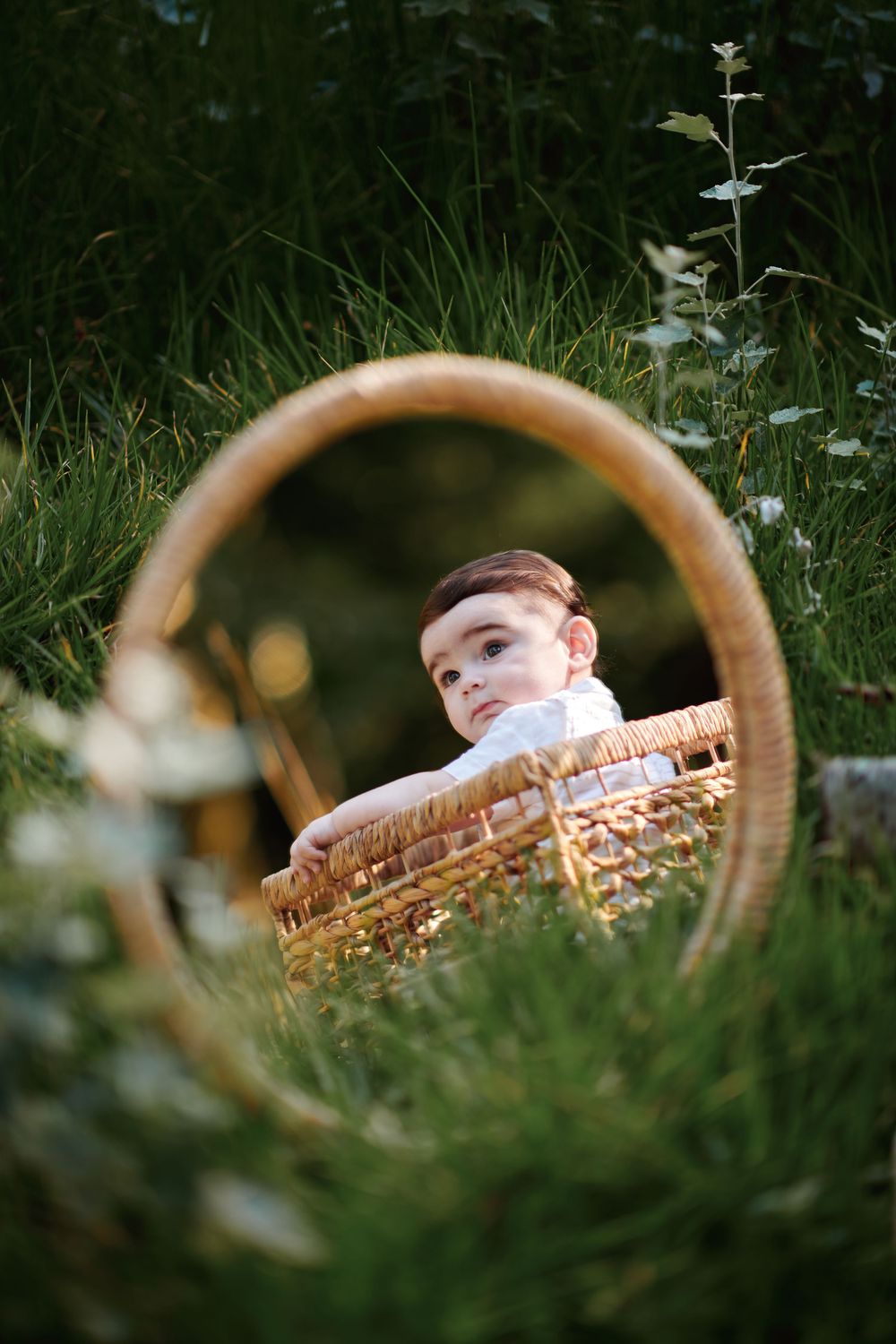 A curious baby peeks over the edge of a large wicker basket while sitting in a lush green garden setting.
