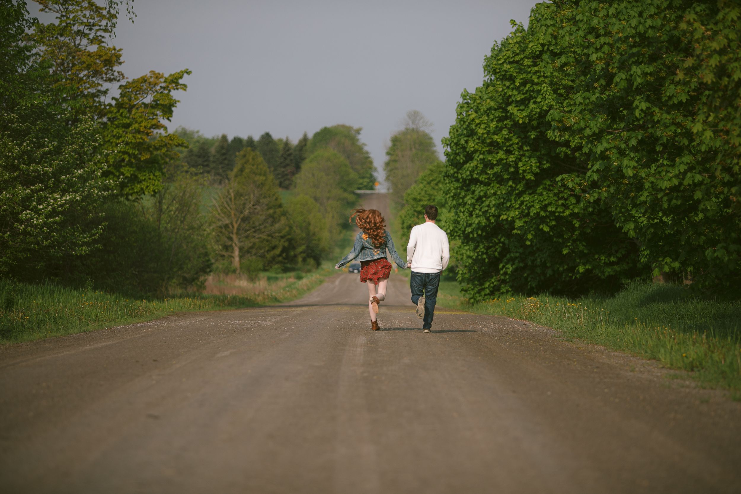 A couple strolls together on a tree-lined dirt path through lush green countryside on a sunny day.