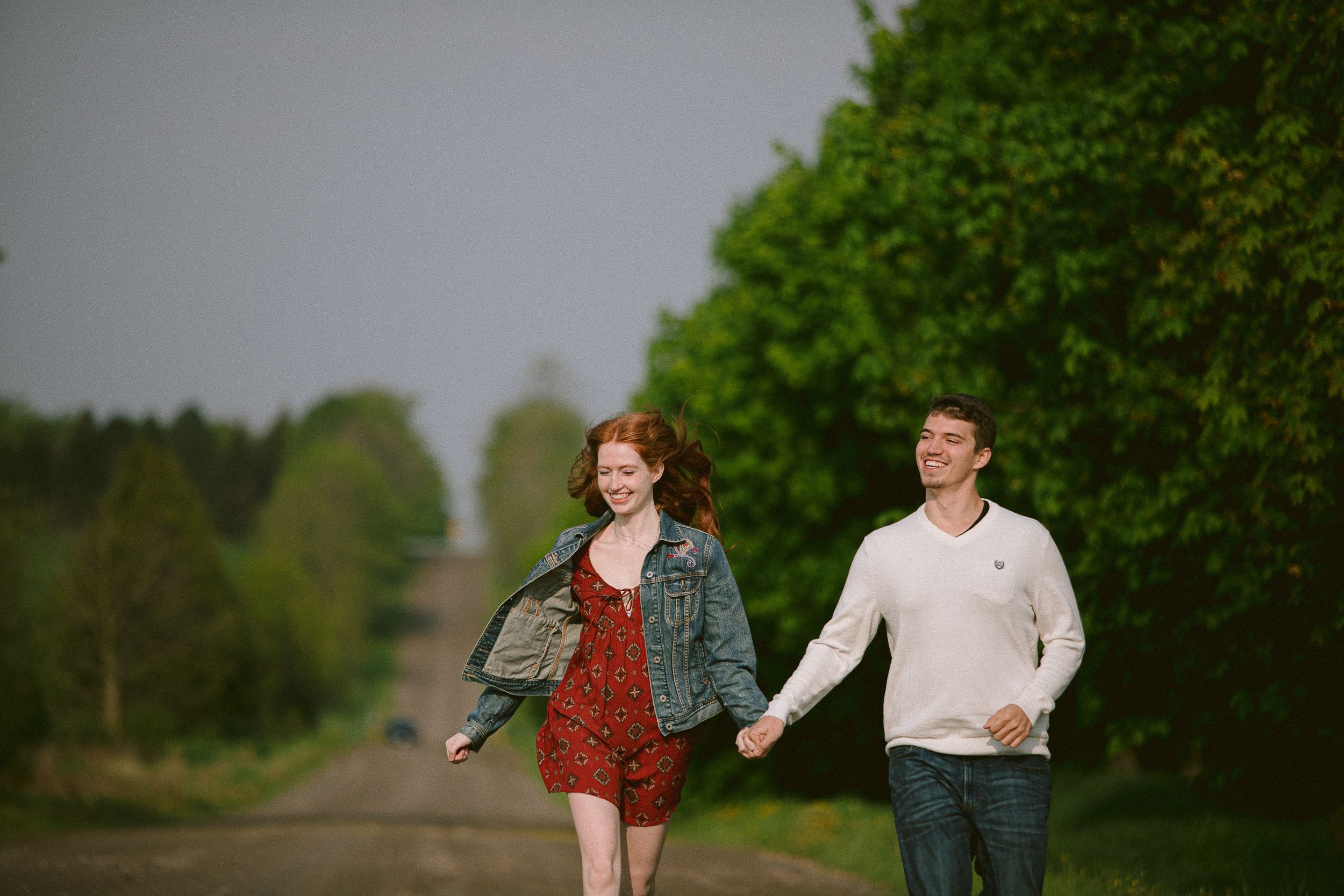 Two people walk hand in hand down a scenic rural path surrounded by green trees and vegetation.