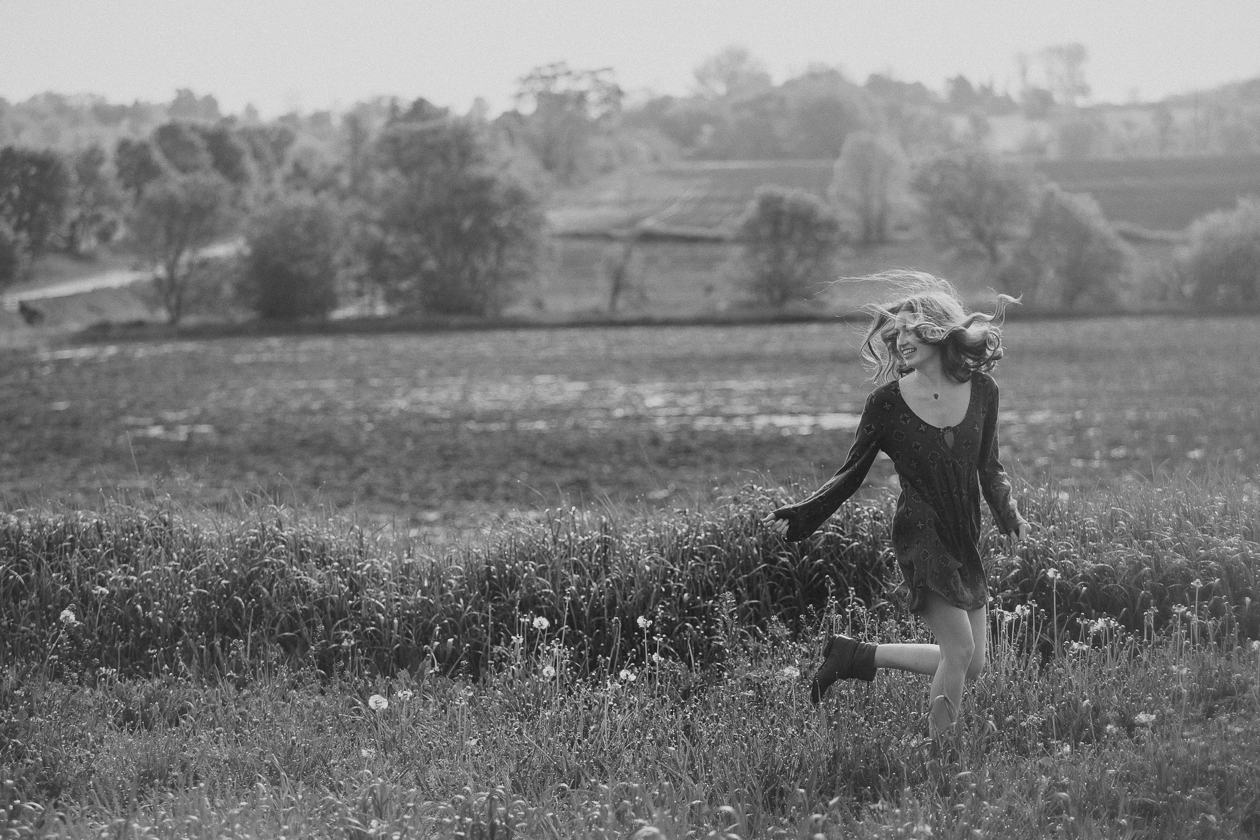 A figure in vintage dress stands in a dreamy black and white meadow with rolling hills and trees in the background.