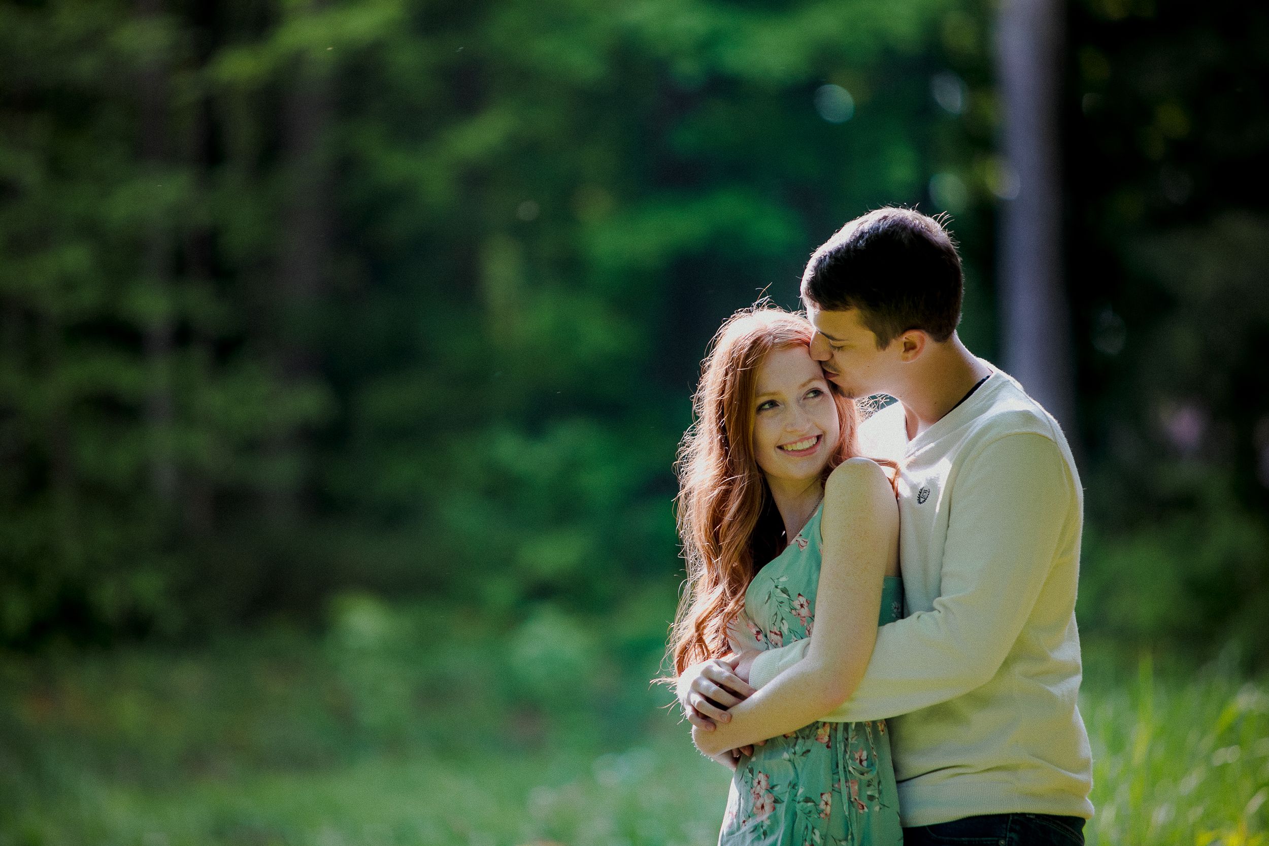 A romantic couple embraces outdoors with soft focus green forest backdrop.
