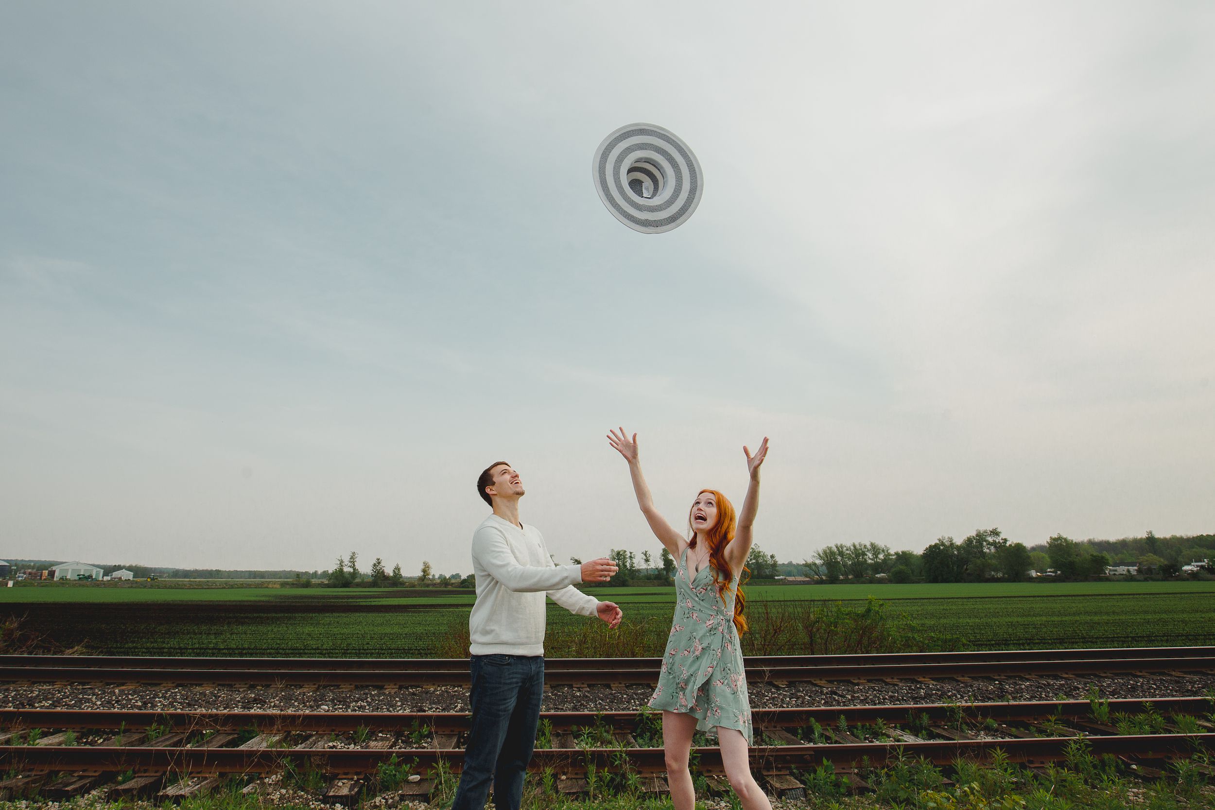 A couple plays with a flying disc by railroad tracks in a playful engagement photo.