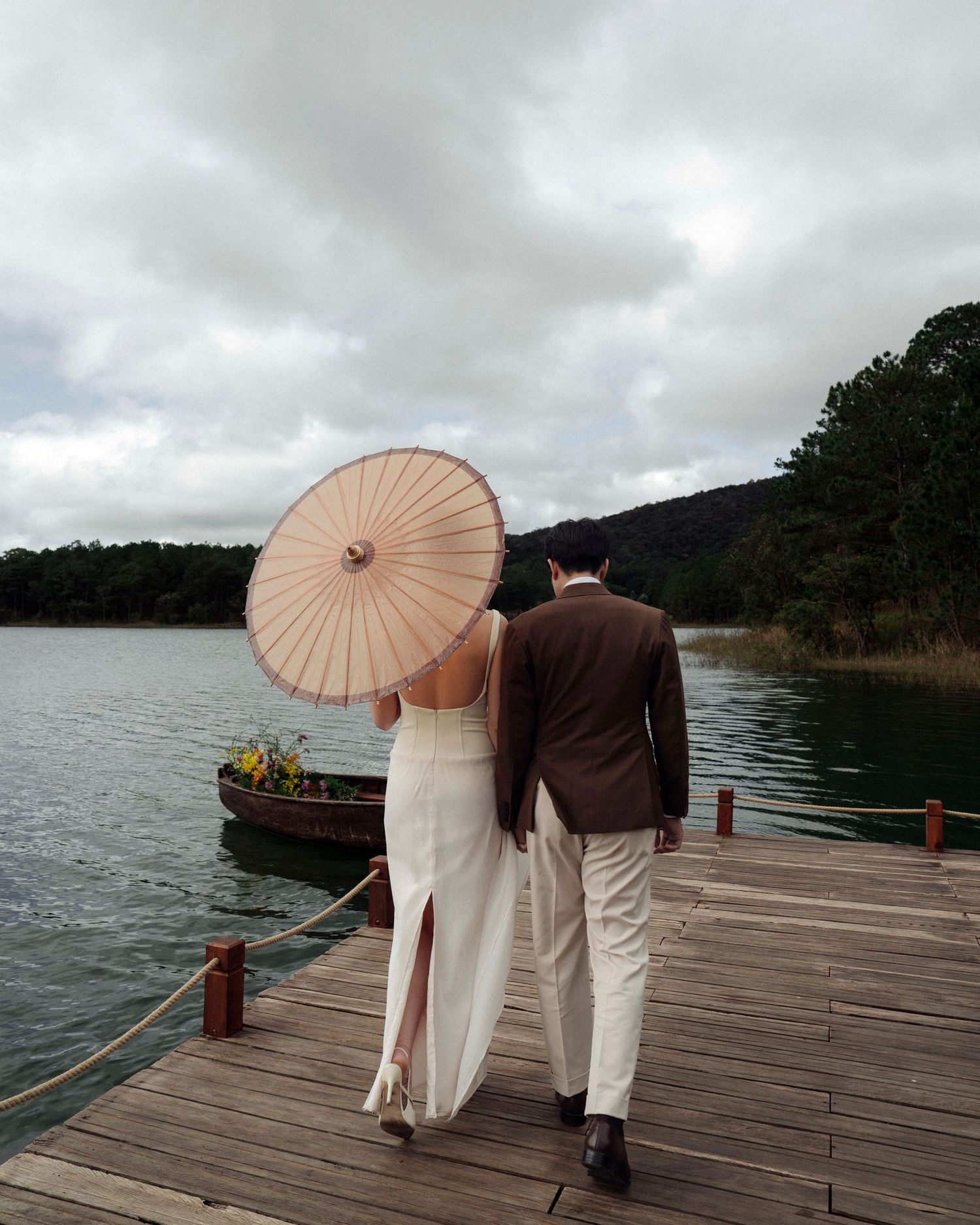 Couple walking together on a lakeside dock, bride in white dress holding a parasol, groom in brown suit.