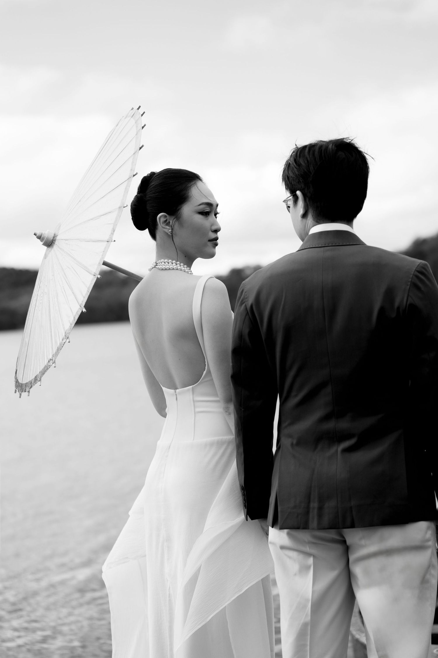 Bride in backless white gown holding parasol stands with groom by waterfront in black and white.