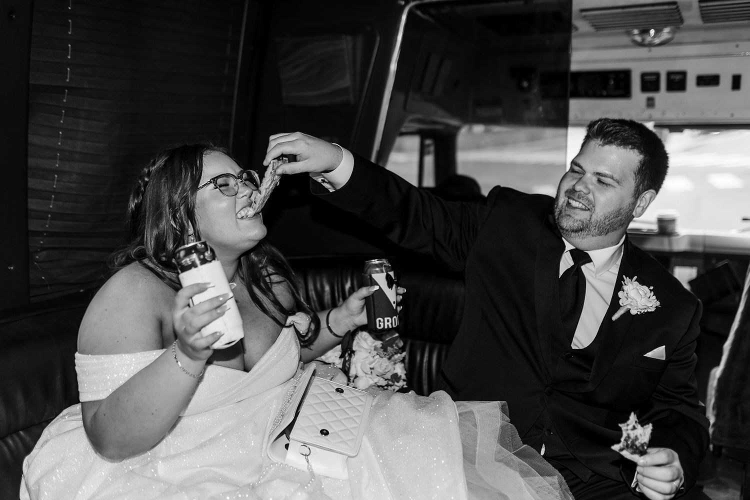 A couple in wedding attire share a playful moment while enjoying drinks in a black and white photograph.