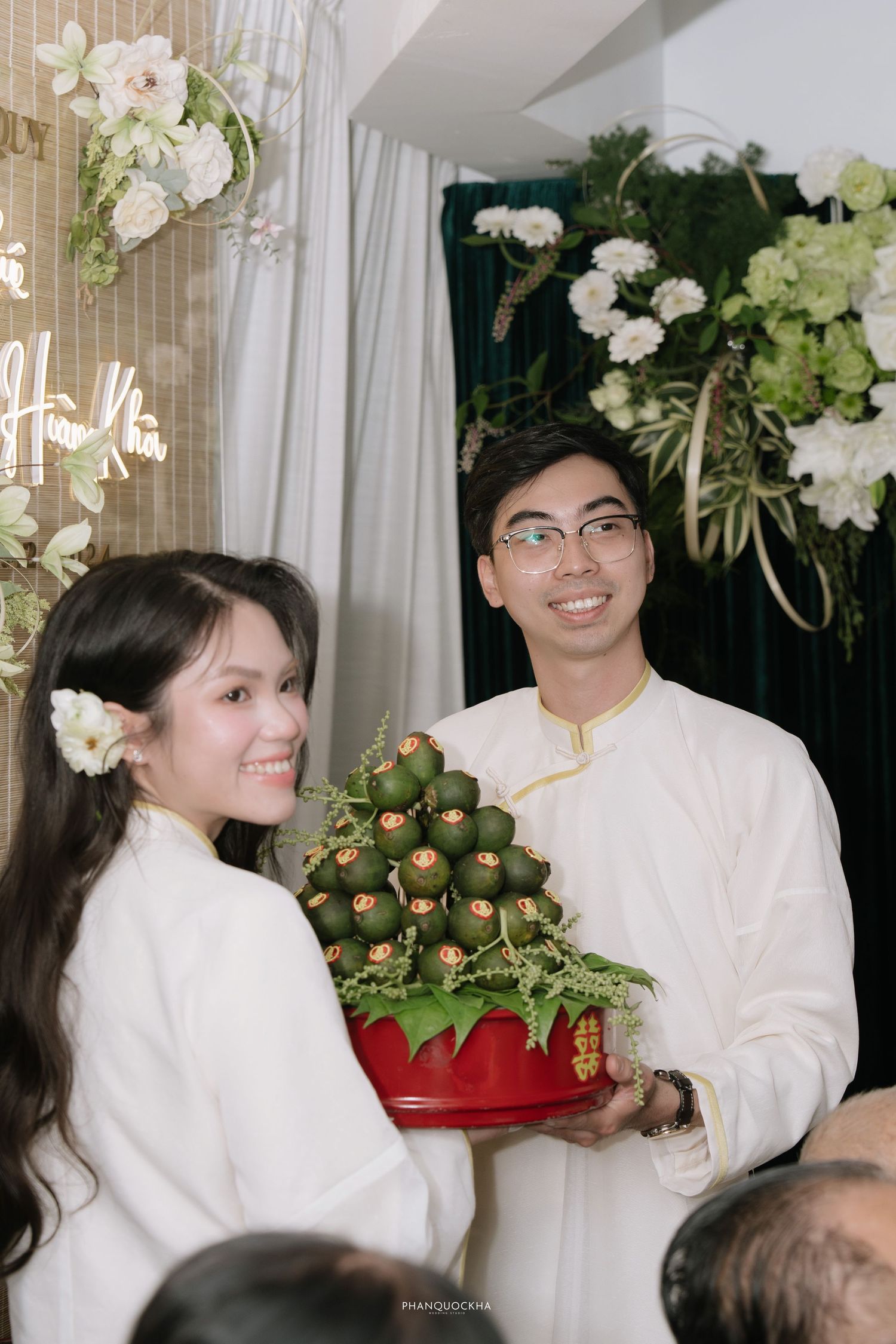 Smiling Vietnamese couple in white ao dai holding traditional betel nut arrangement at engagement ceremony.