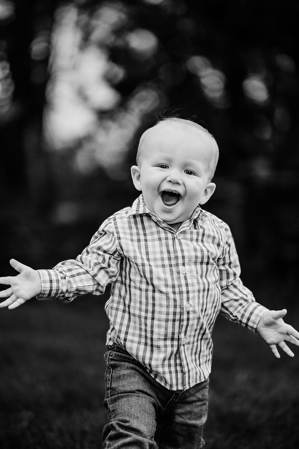 A young child in a plaid shirt and jeans expresses pure joy with arms outstretched outdoors in black and white.