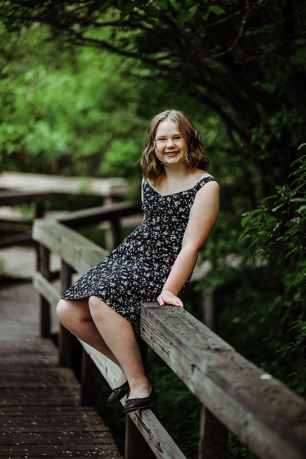 A person in a floral print dress sits on a wooden bridge railing surrounded by lush green forest.