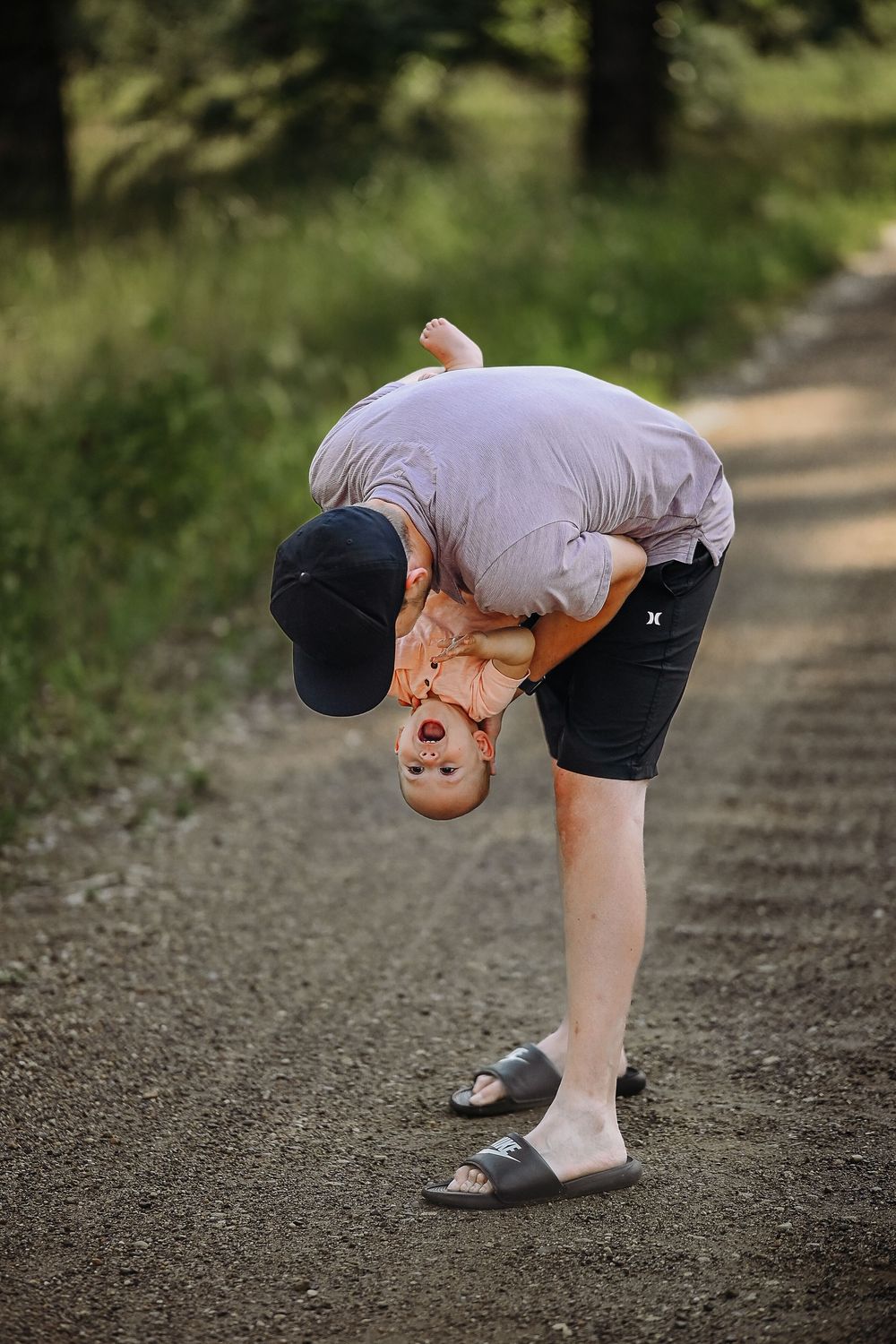 Person in black baseball cap and shorts bends down to look at something on a dirt path in a natural setting.