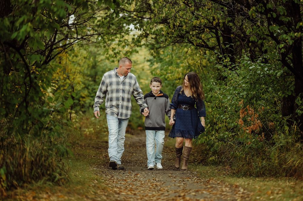 A family walks together on a tree-lined path during autumn wearing casual clothing and plaid shirts.