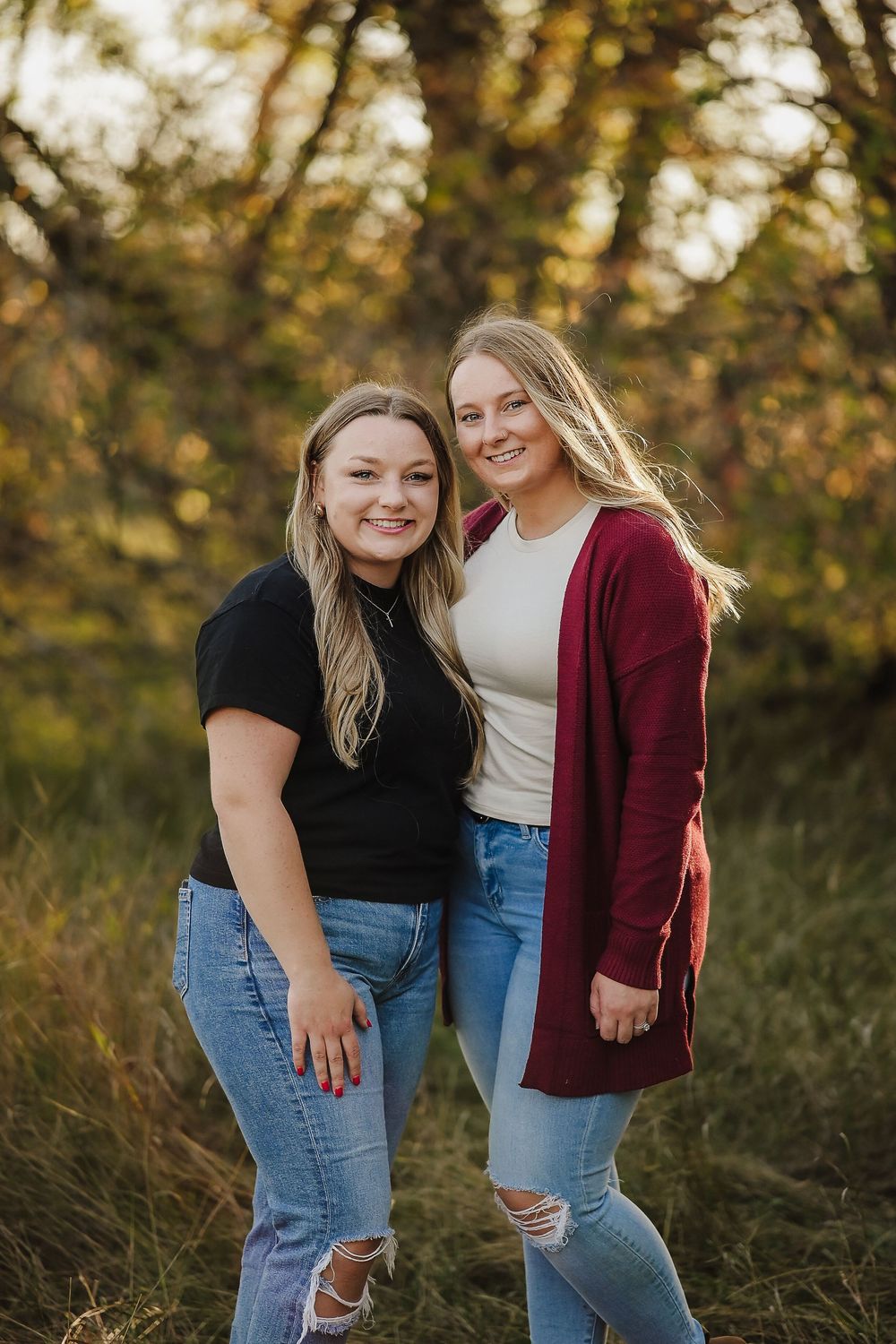 Two friends in matching jeans pose playfully outdoors during golden hour with autumn foliage in the background.