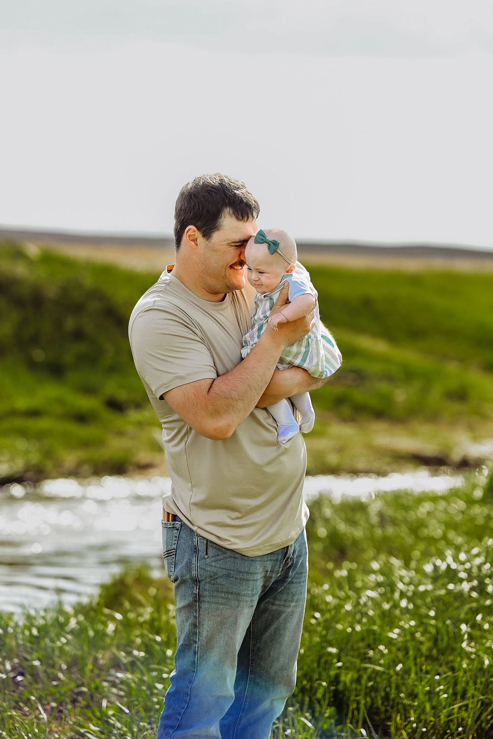 Parent and baby share loving moments in field of white flowers near water on sunny afternoon.