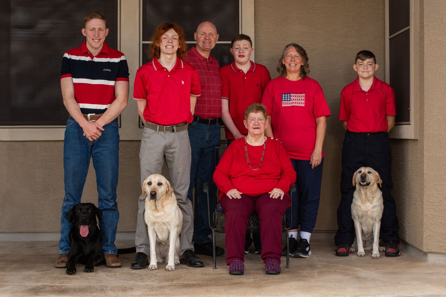 A group in coordinated red shirts pose together on a porch with three well-behaved dogs.