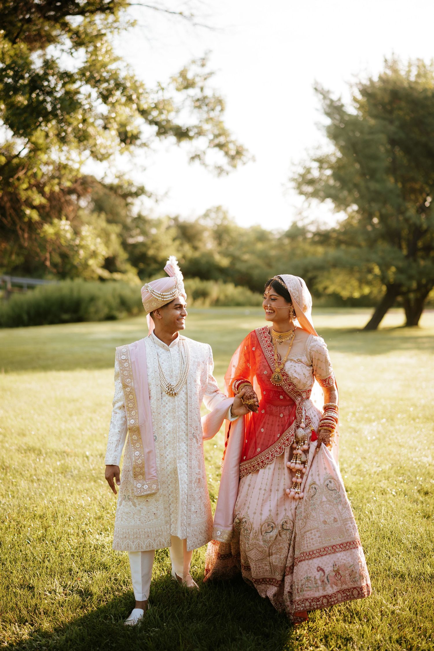 A couple in traditional South Asian wedding attire walk together on a sunlit grassy field during golden hour.