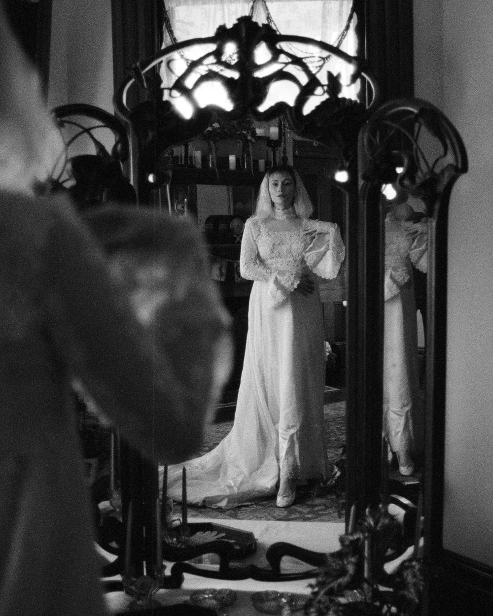 Black and white photo of a person in a white dress reflected in an ornate vintage mirror with decorative metalwork frame.
