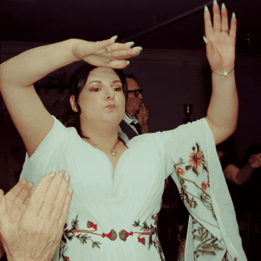 Someone in a white embroidered dress with floral details dances with raised arms at a celebration.