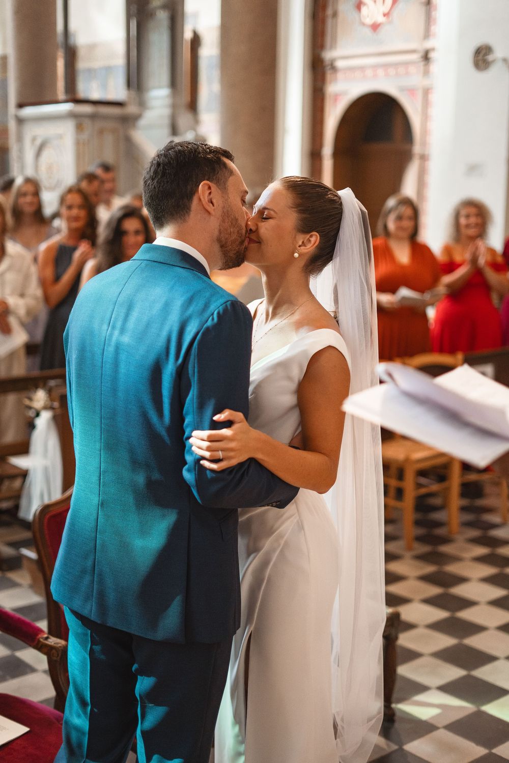Newlyweds share a kiss in a church with black and white checkered floors and guests watching from the pews.