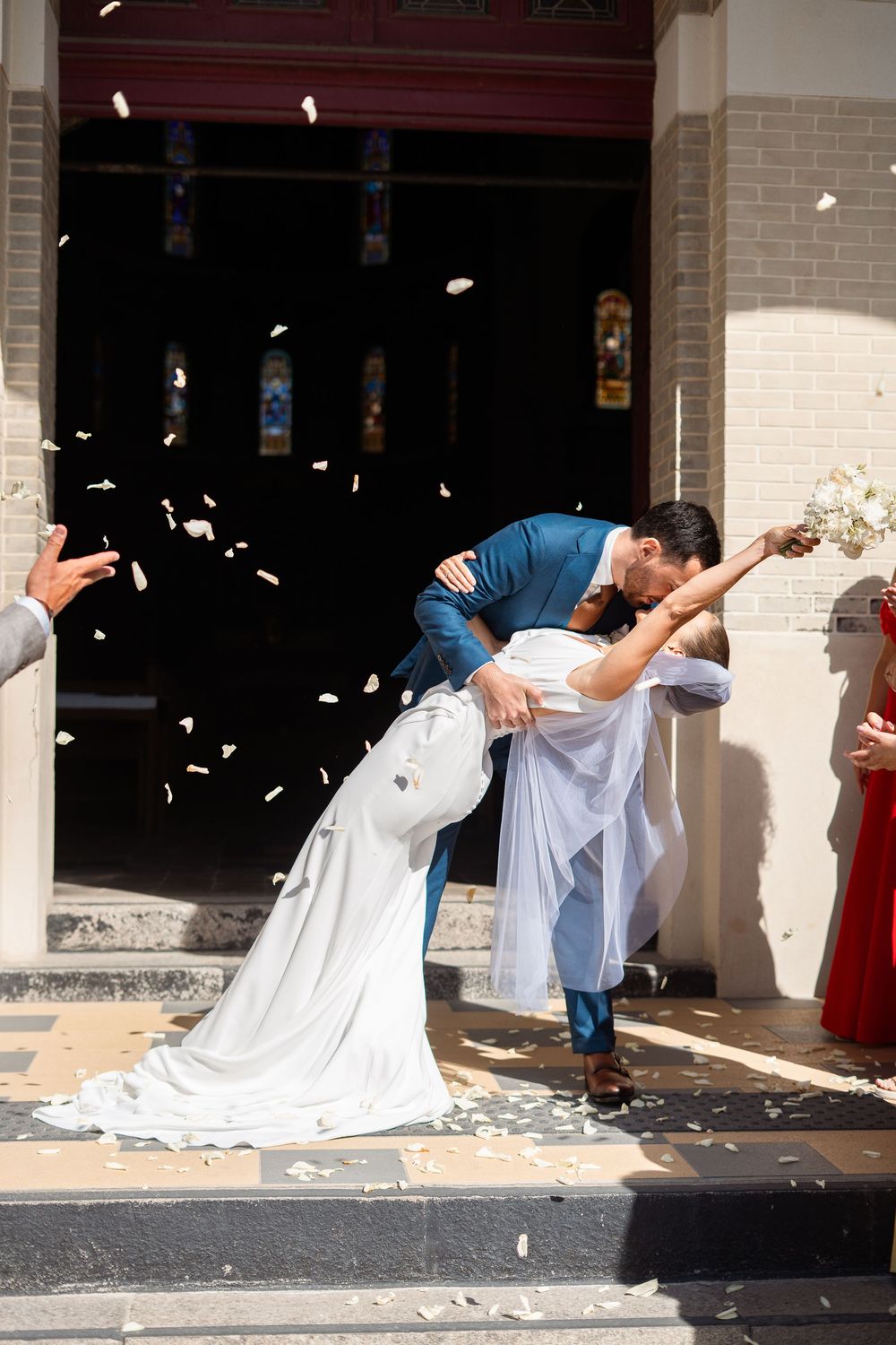 A romantic couple shares a kiss on church steps while rose petals fall around them after their wedding ceremony.