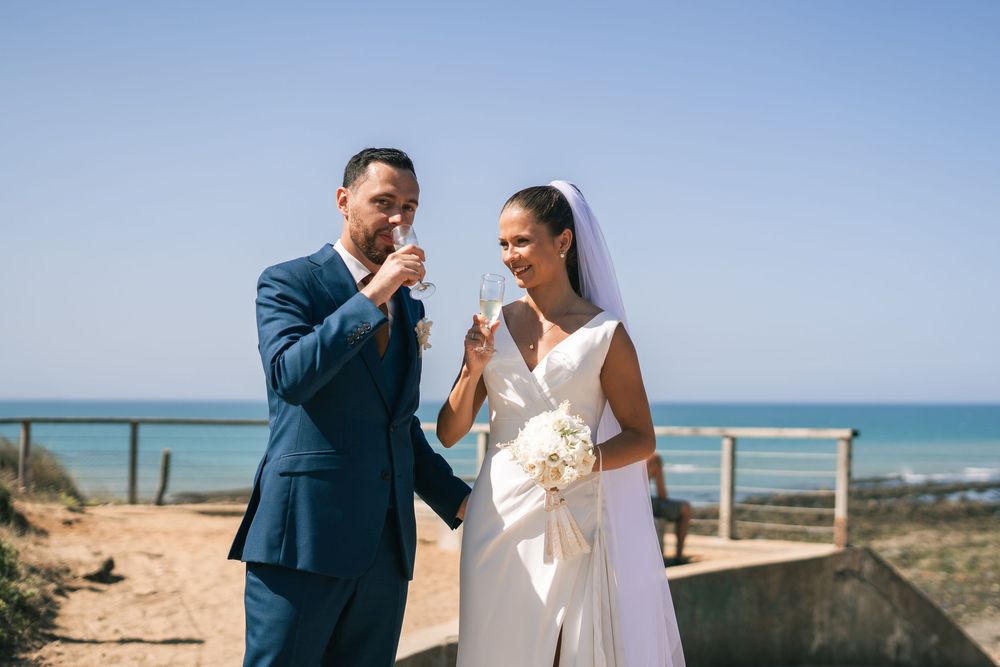 Elegant wedding couple poses together by the ocean on a bright sunny day with sandy beach and blue water backdrop.
