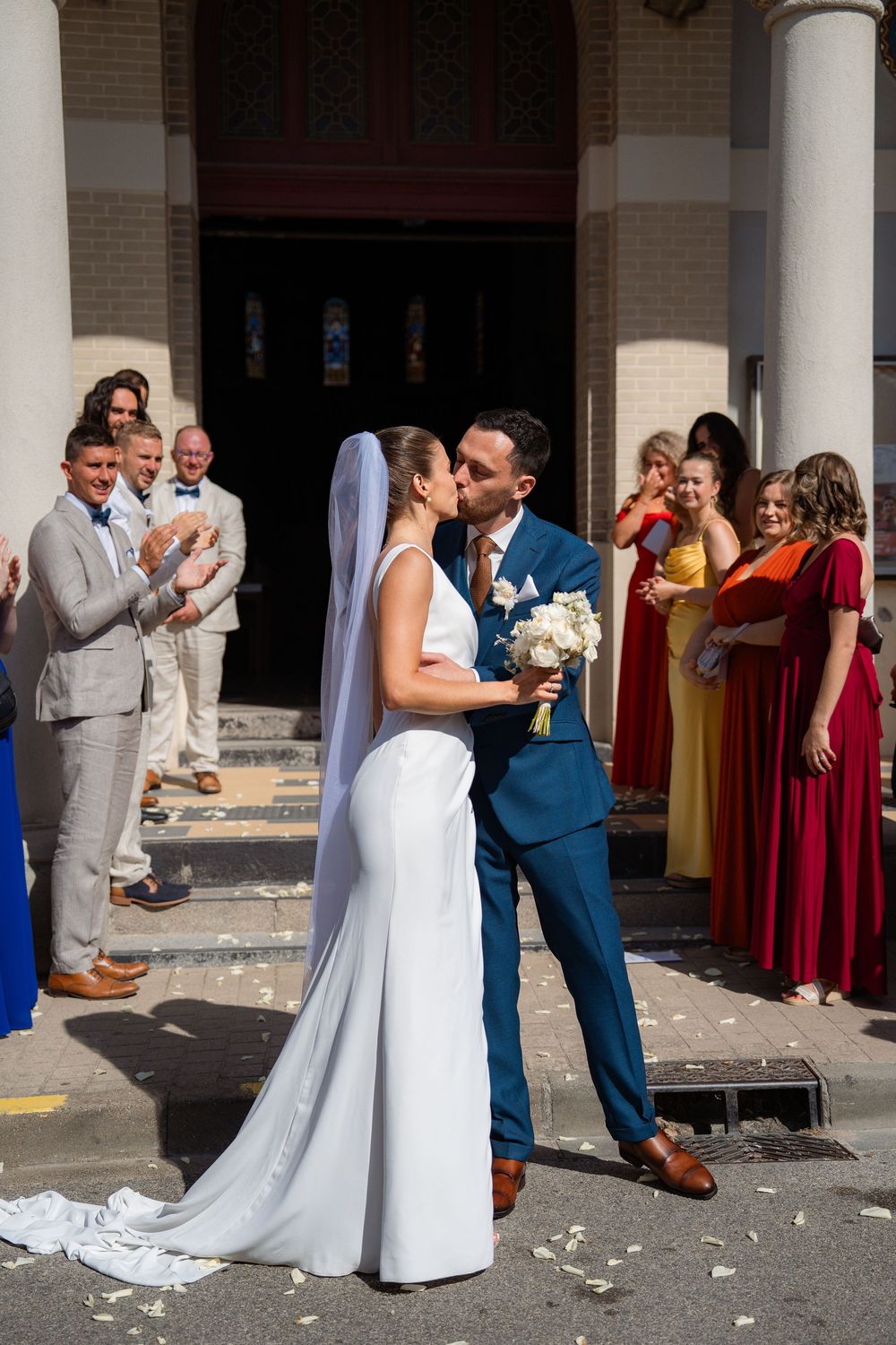 Wedding celebration outside church with bridal party in burgundy and blue attire throwing confetti.