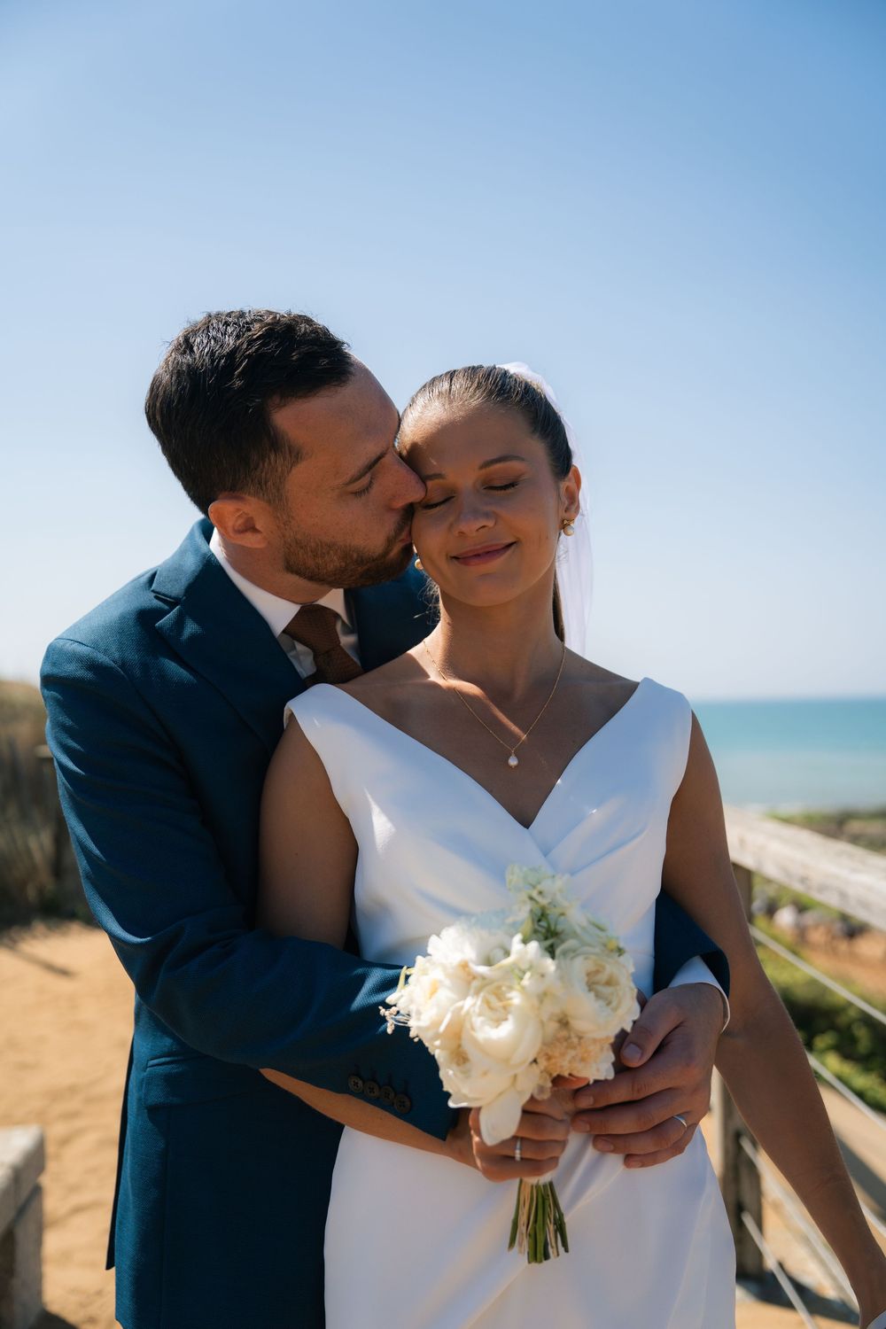 A couple shares tender moments on their wedding day against a bright blue coastal backdrop.