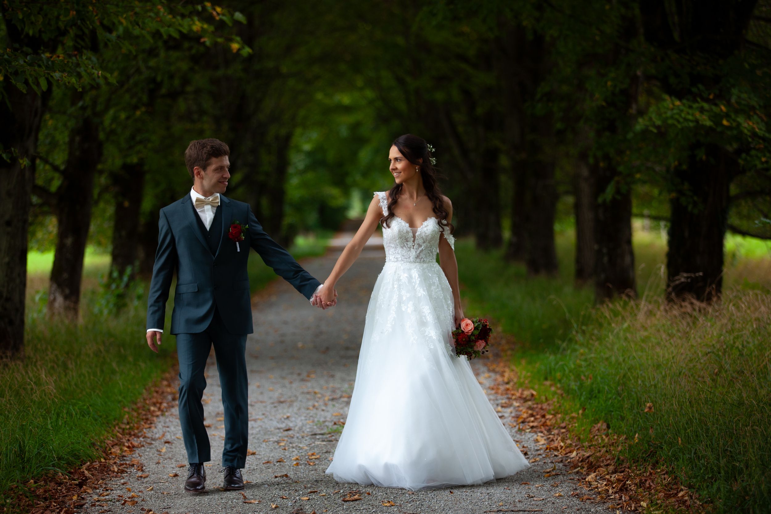 Newlyweds walk hand in hand down a tree-lined path in an autumn park setting.