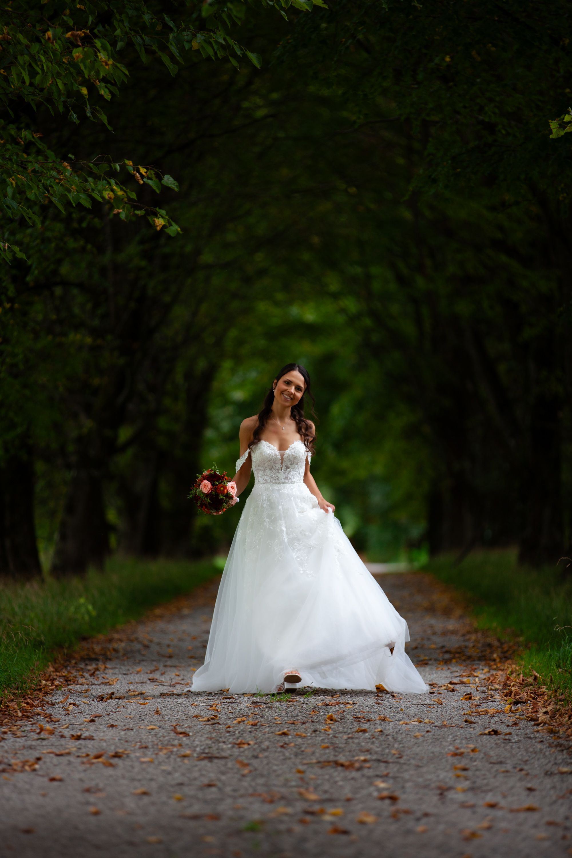 A romantic forest path portrait of someone in a flowing white wedding gown among autumn leaves and greenery.
