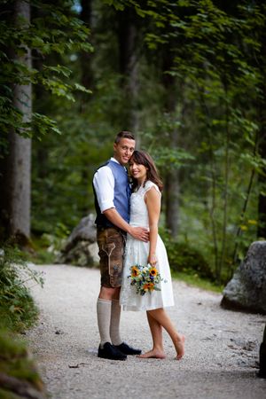A romantic couple walking on a forest path together wearing Bavarian-inspired attire with a colorful bouquet.