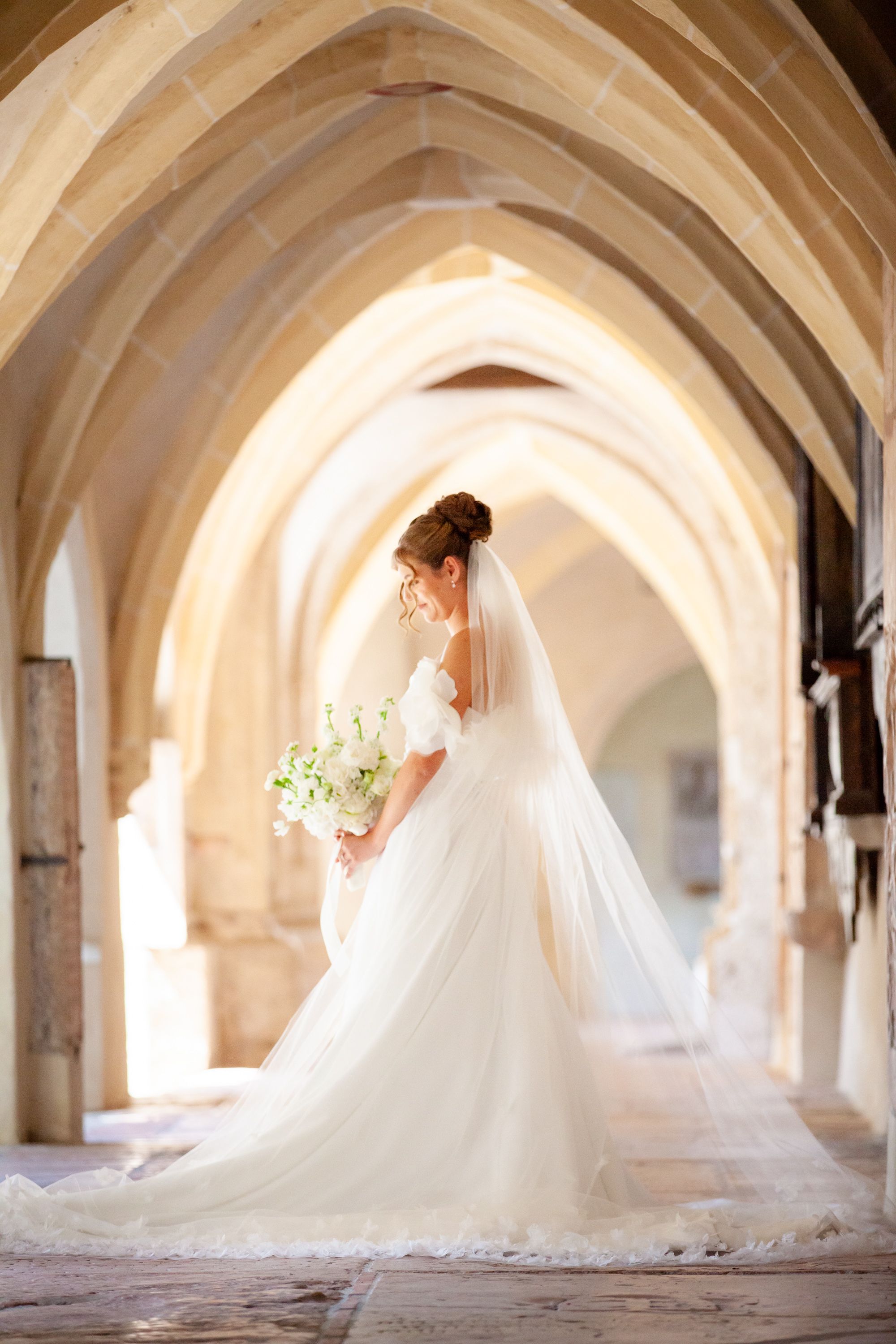 Bride in white gown and veil holding bouquet stands under Gothic stone arches in elegant cathedral corridor.