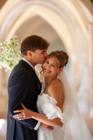 Groom in navy suit kissing bride in white off-shoulder gown, smiling in elegant church with arched stone walls.