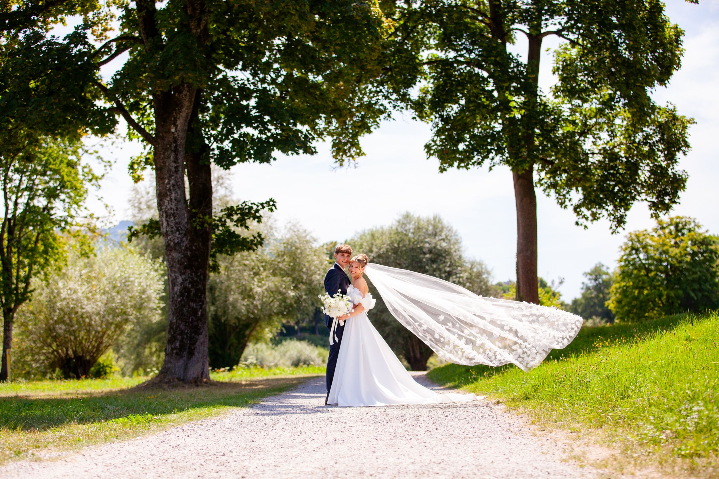 Bride and groom kissing on a gravel path surrounded by lush green trees, with the wedding veil flowing in the wind.