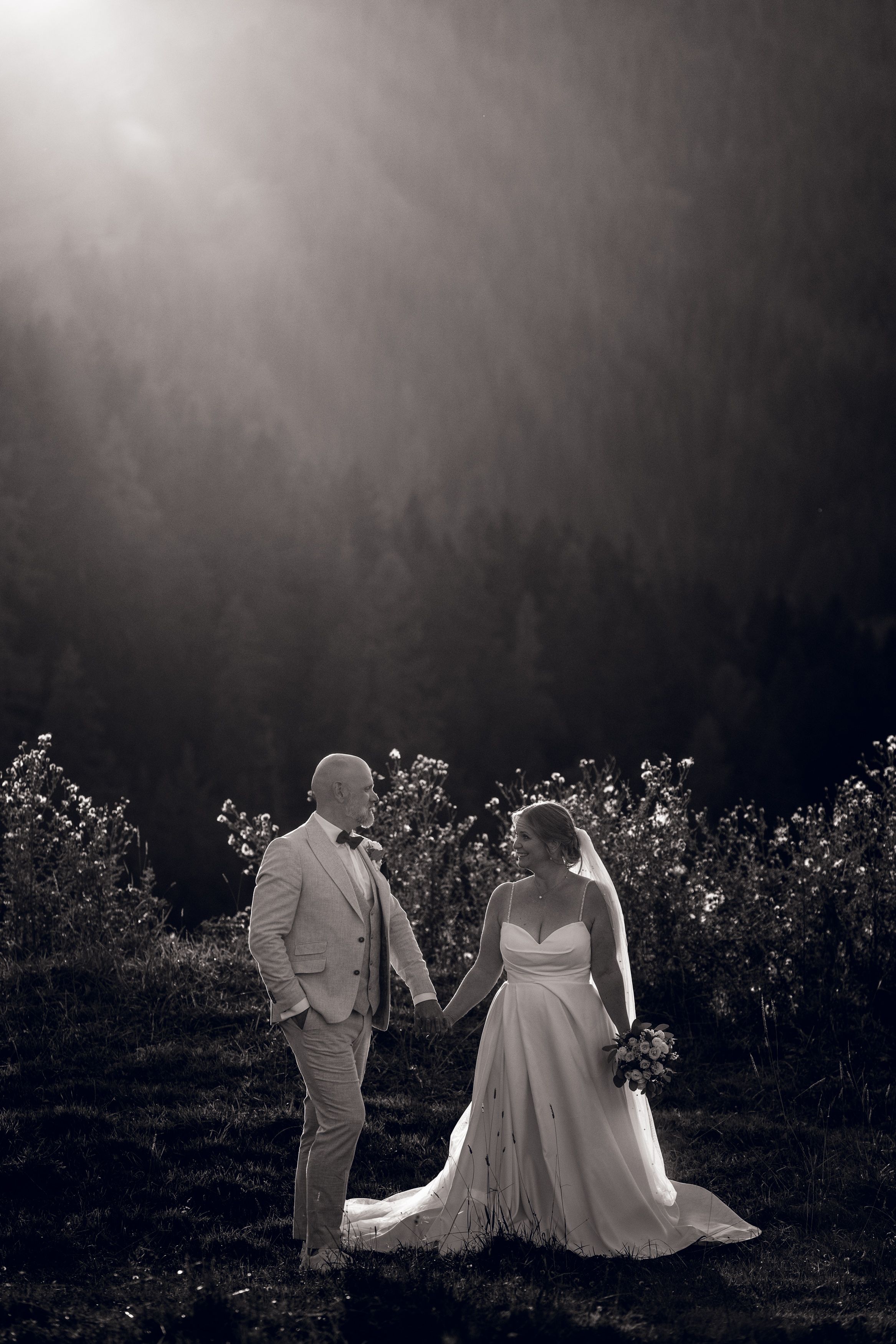 Black and white wedding photo of bride and groom holding hands in a sunlit field with forest backdrop.
