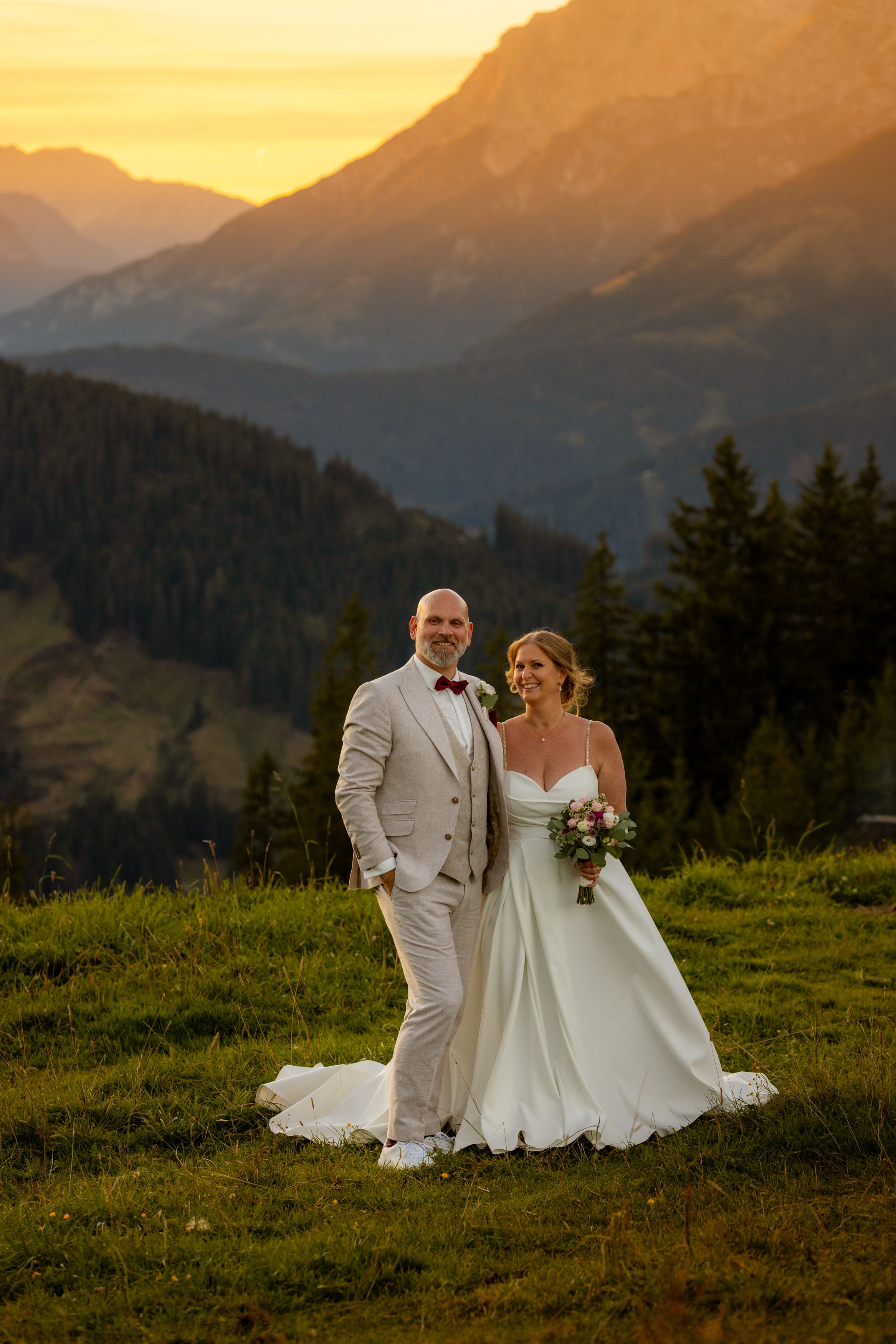 Bride and groom pose at golden sunset in Alpine meadow, she holds bouquet in white gown, he wears beige suit with red bow tie.