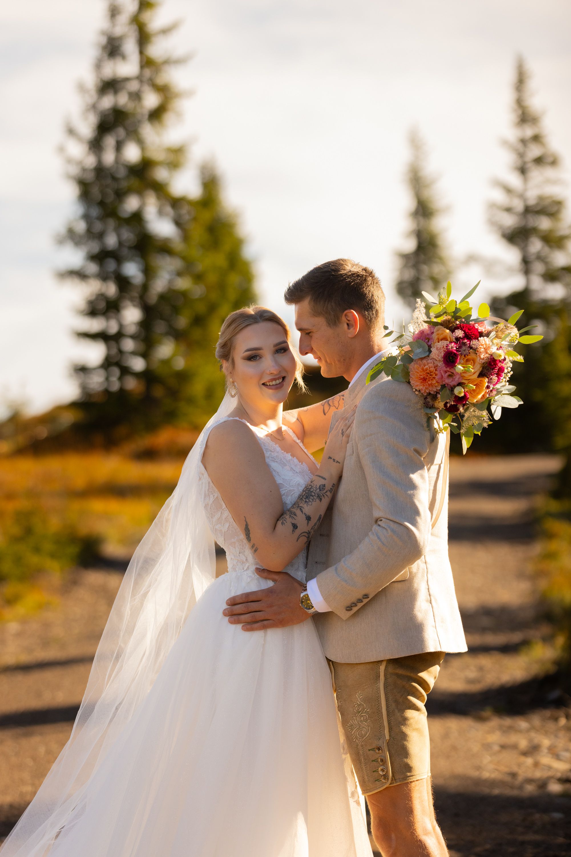 Bride and groom embracing outdoors with evergreen trees, holding a colorful bouquet, in golden hour light.