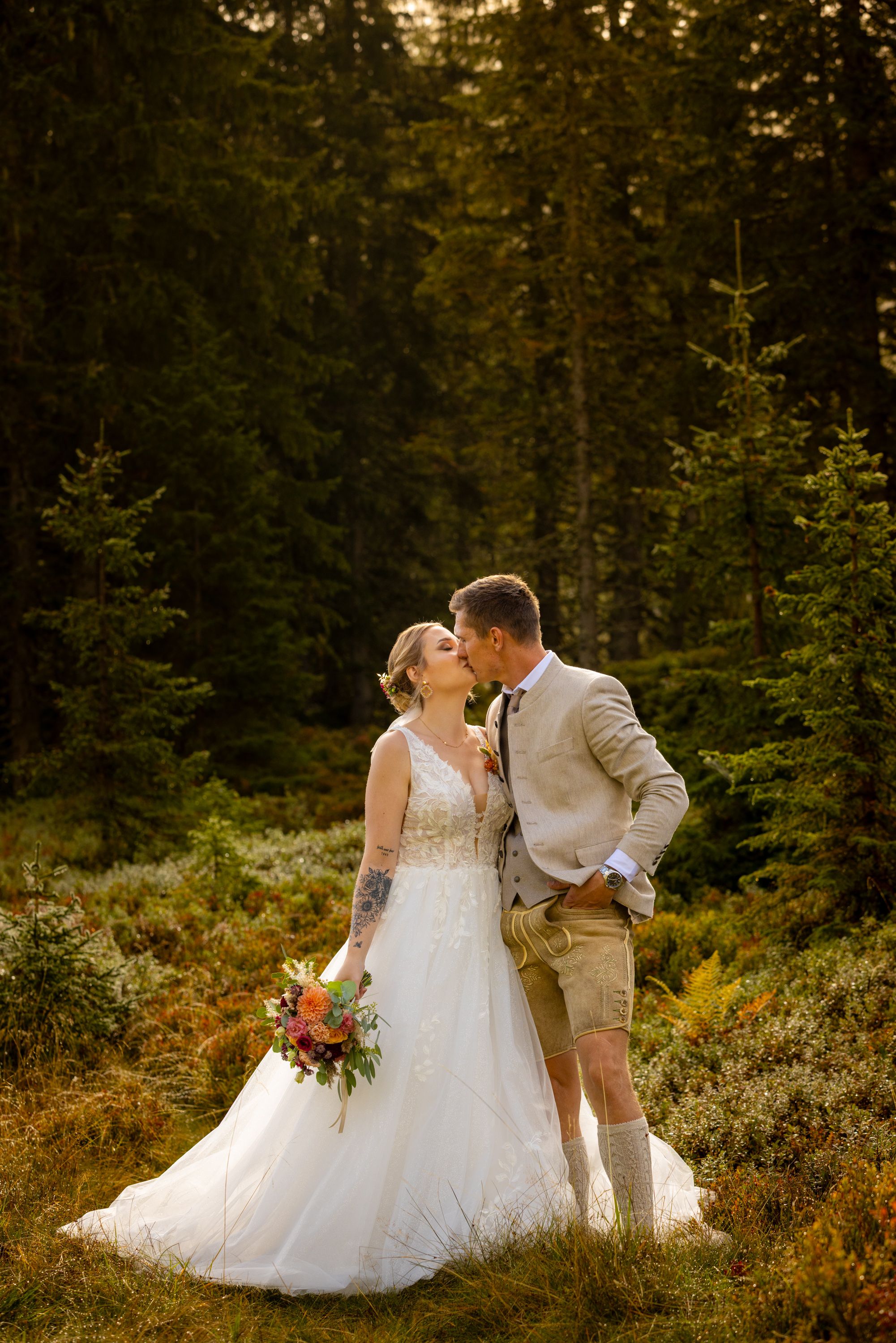 Bride and groom share a romantic kiss in a sunlit forest clearing, bride holding a colorful bouquet in a white gown.