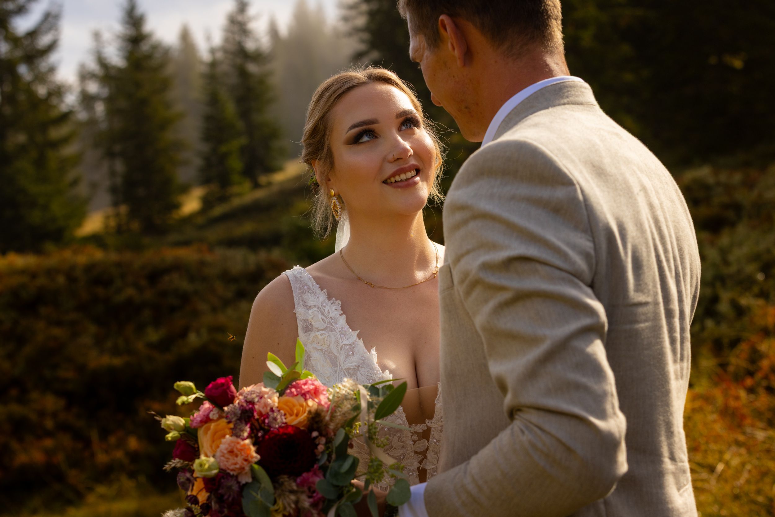 Bride smiling at groom outdoors in golden light, holding colorful bouquet, wearing lace dress, groom in tan suit, forest backdrop.