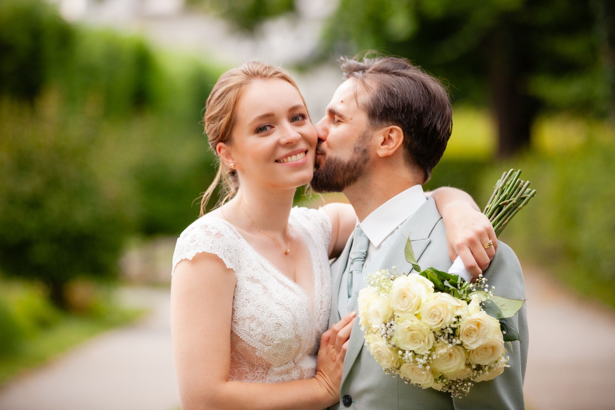 A romantic wedding couple embraces outdoors in a garden setting with yellow and white bouquet.