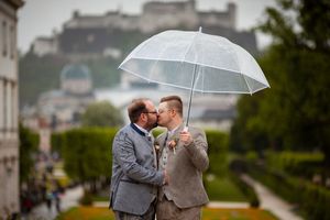 Two people share a romantic moment under a clear umbrella in a rainy European city setting.