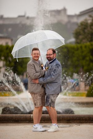 Two people holding clear umbrellas share a joyful moment near a fountain on a cloudy day.