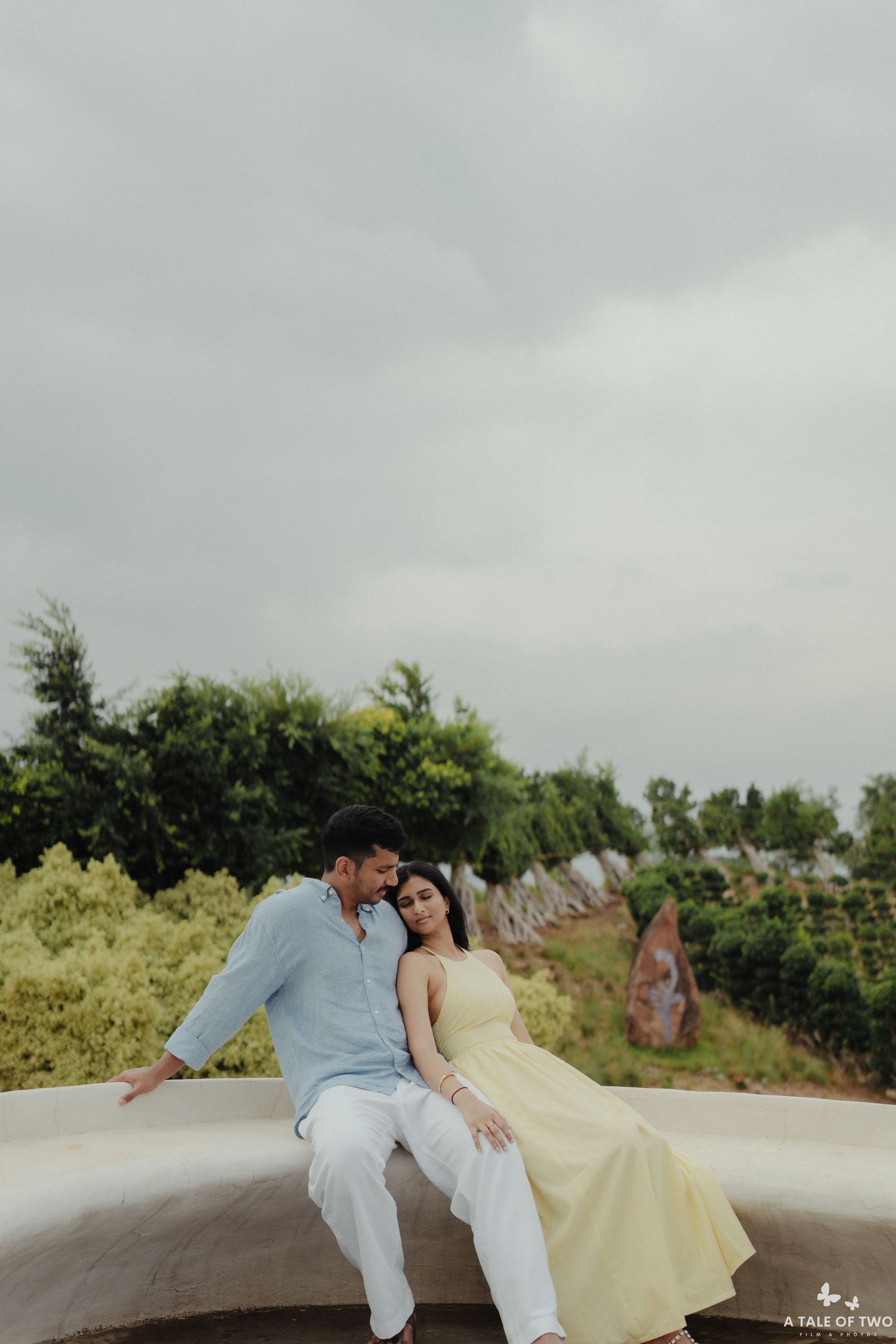 A couple in white and light blue outfits share a romantic moment while sitting on a wall with greenery in the background.