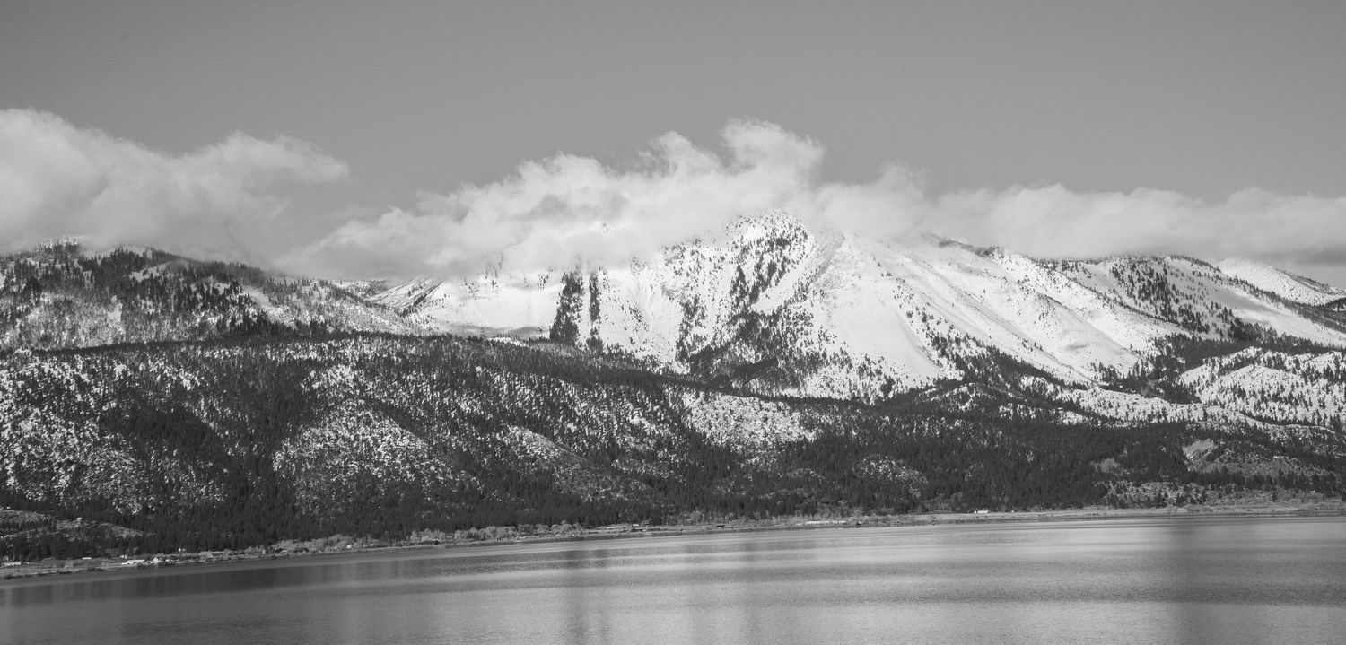 Black and white panoramic view of snow-capped mountains reflecting over a calm lake under cloudy skies.
