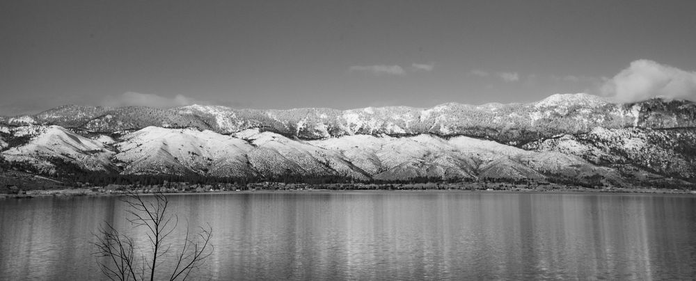 Black and white panoramic view of snow-capped mountains reflecting in a calm lake with bare trees in foreground.