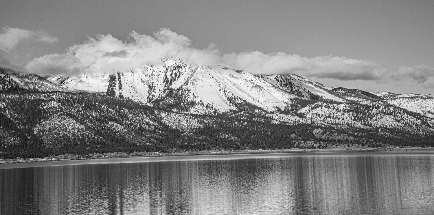 Black and white panoramic photo of snow-capped mountains reflecting on a calm lake with pine trees along the shore.