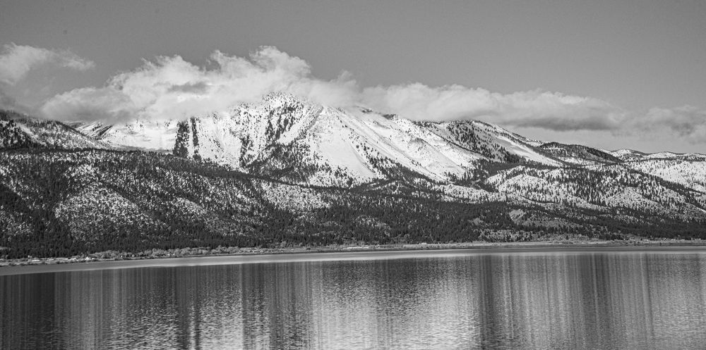 Black and white panoramic photo of snow-capped mountains reflecting on a calm lake with pine trees along the shore.