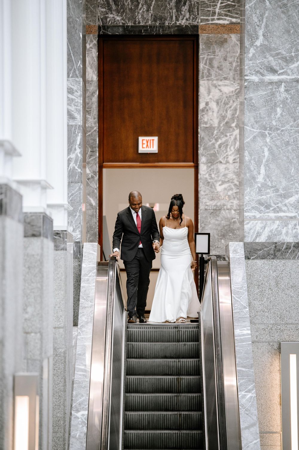 A pair of people descending a marble staircase with stainless steel handrails in a modern building interior.